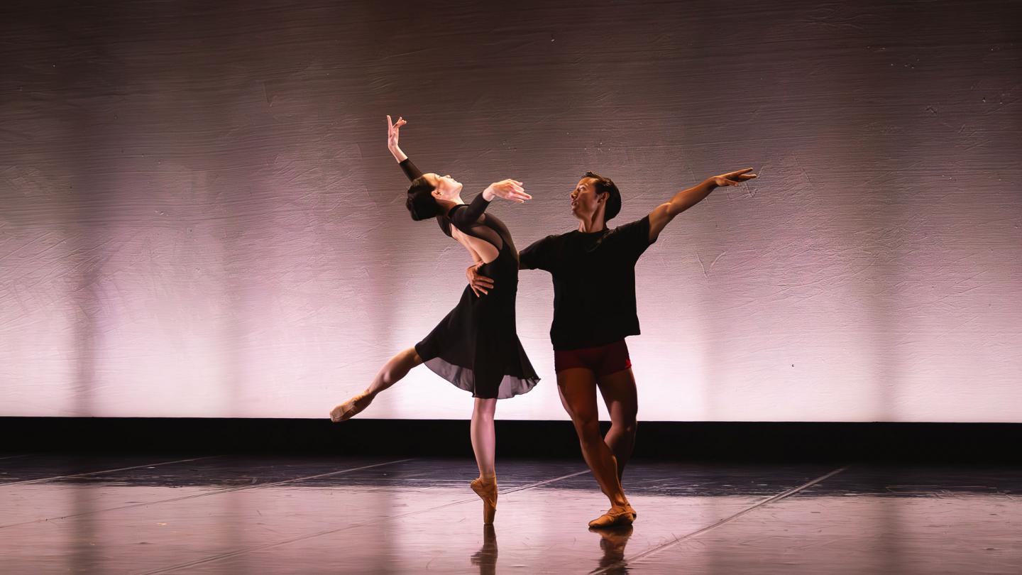 Two dancers stood in a balletic pose against a pink backdrop. 
