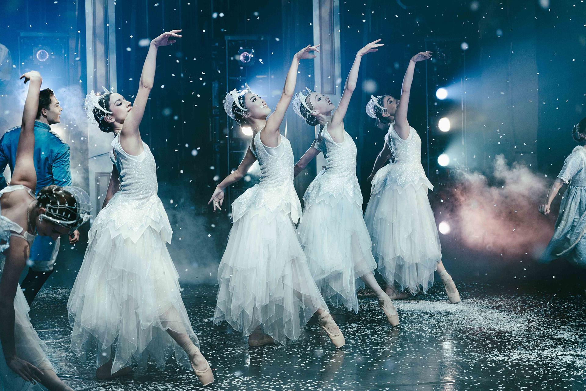 Four female dancers dressed in white each extending a foot down and an arm high