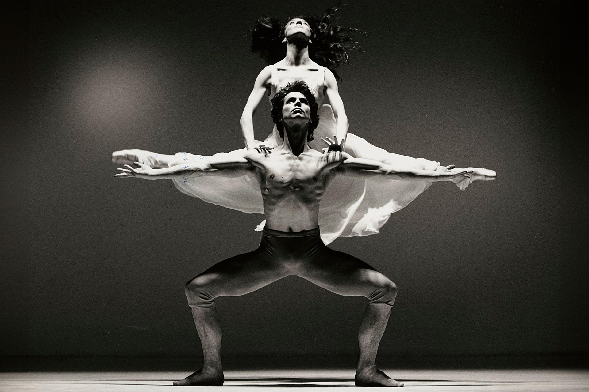 Male dancer, shirtless, standing with his arms wide and a female dancer in a white dress is behind him, holding herself up on his shoulders with her legs split in parallel with the male dancers arms