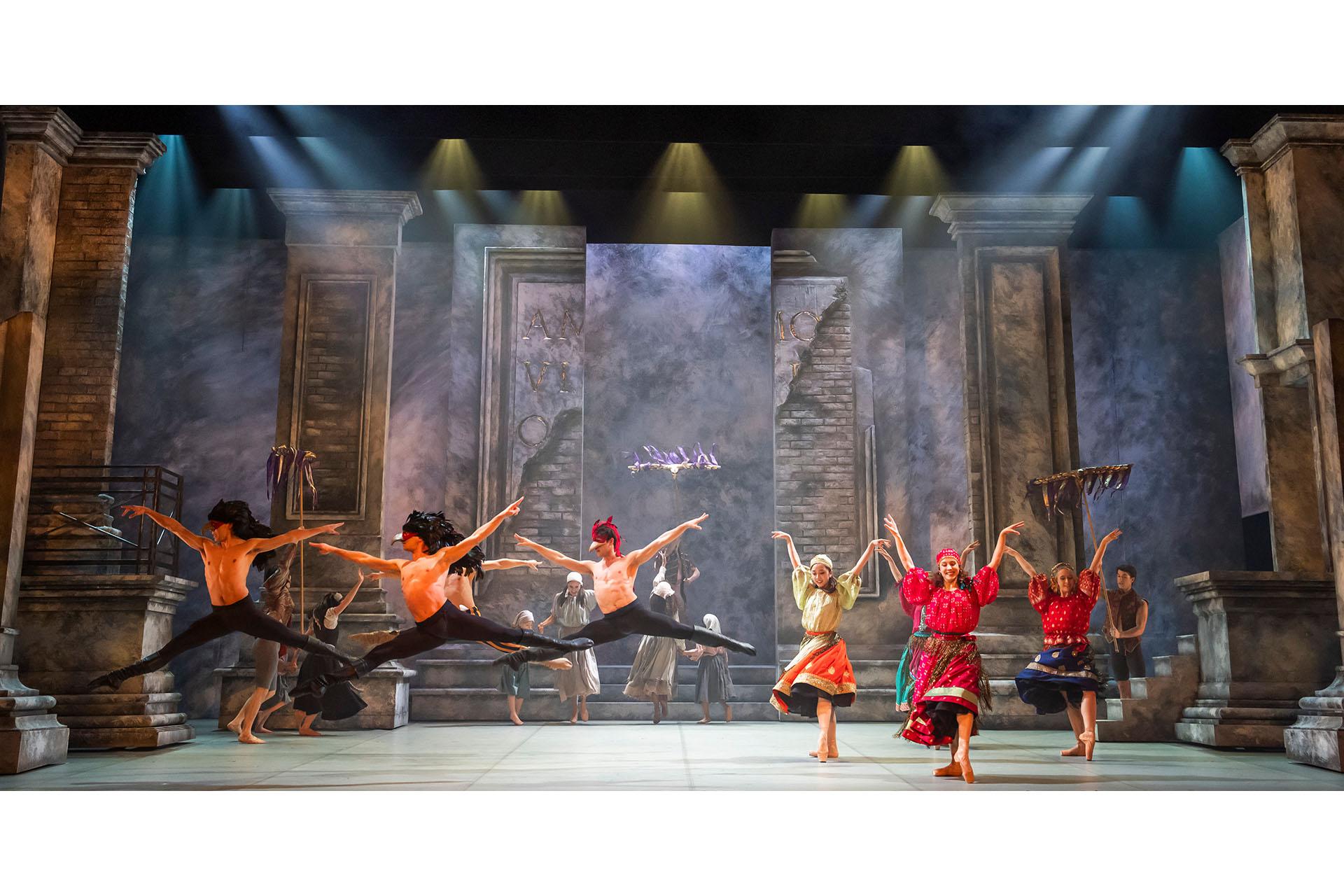 Three dancers on the left of a large stage in bird masks. To the right is a group of ladies in bright skirts.