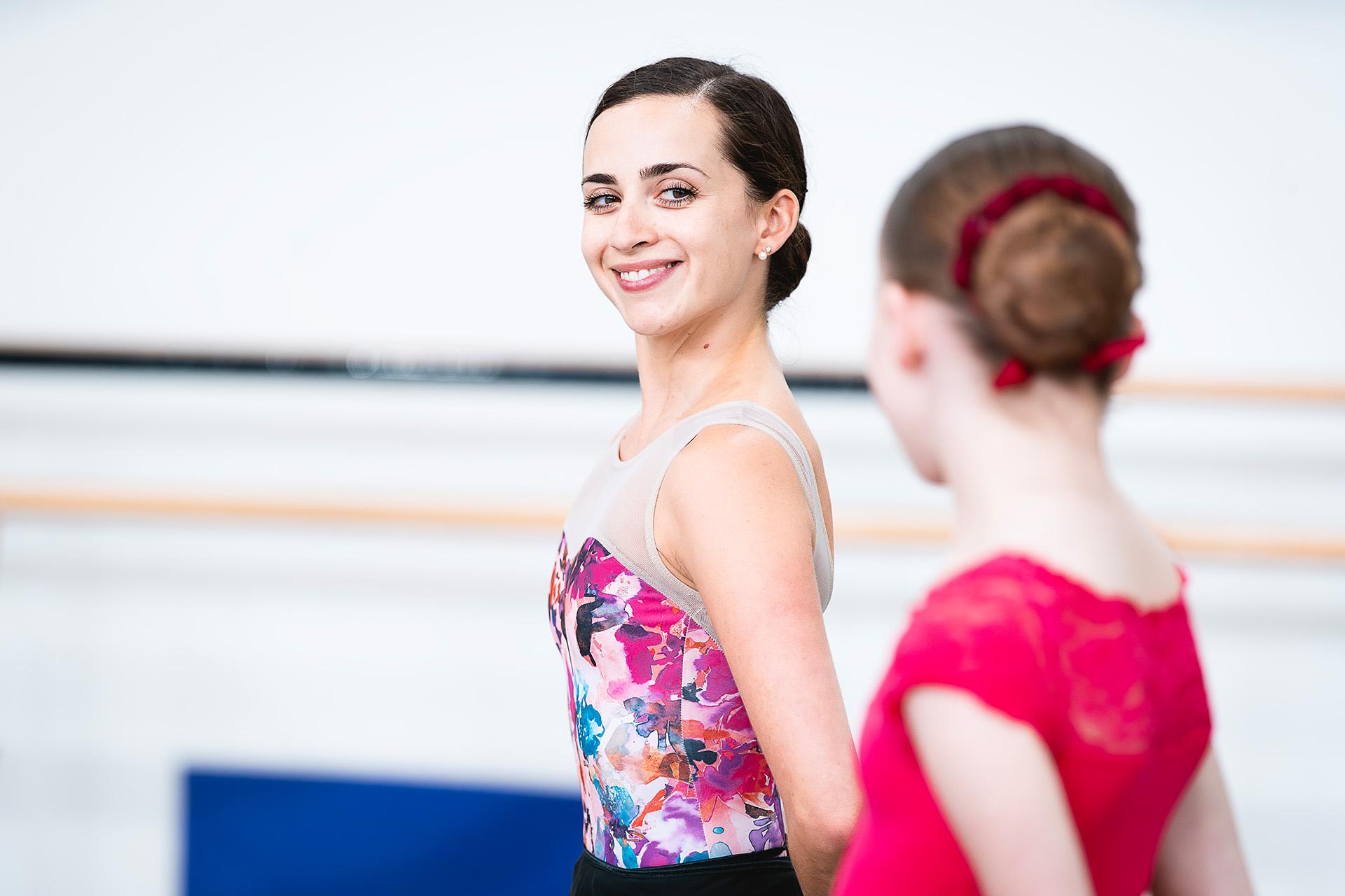 Professional dancer with dark hair tied in a bun stands and smiles encouragingly at a student mirroring her pose