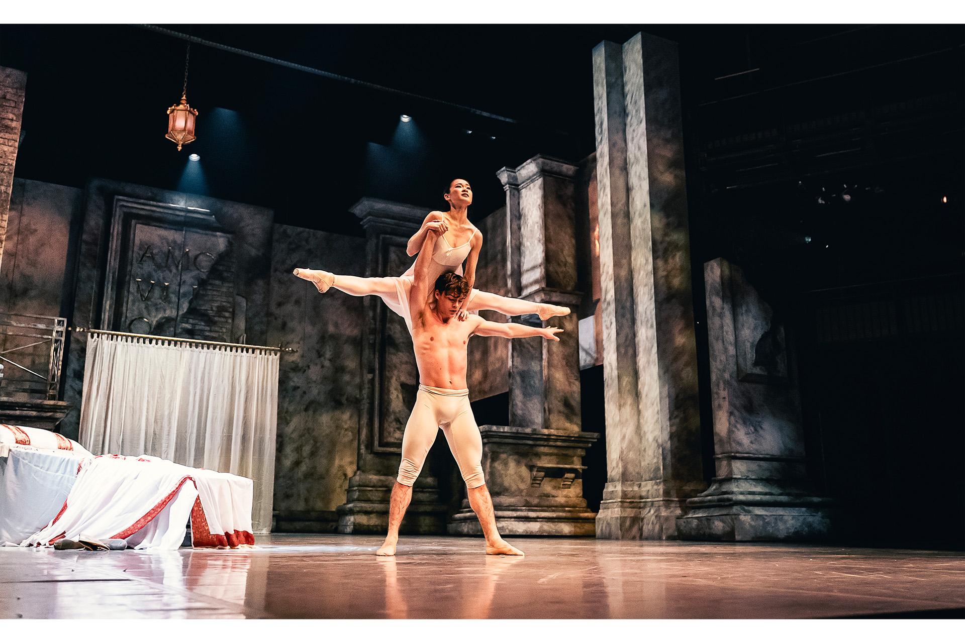 A dancer in white trousers stand in a strong pose, lifting another dancer in a white dress over his head.