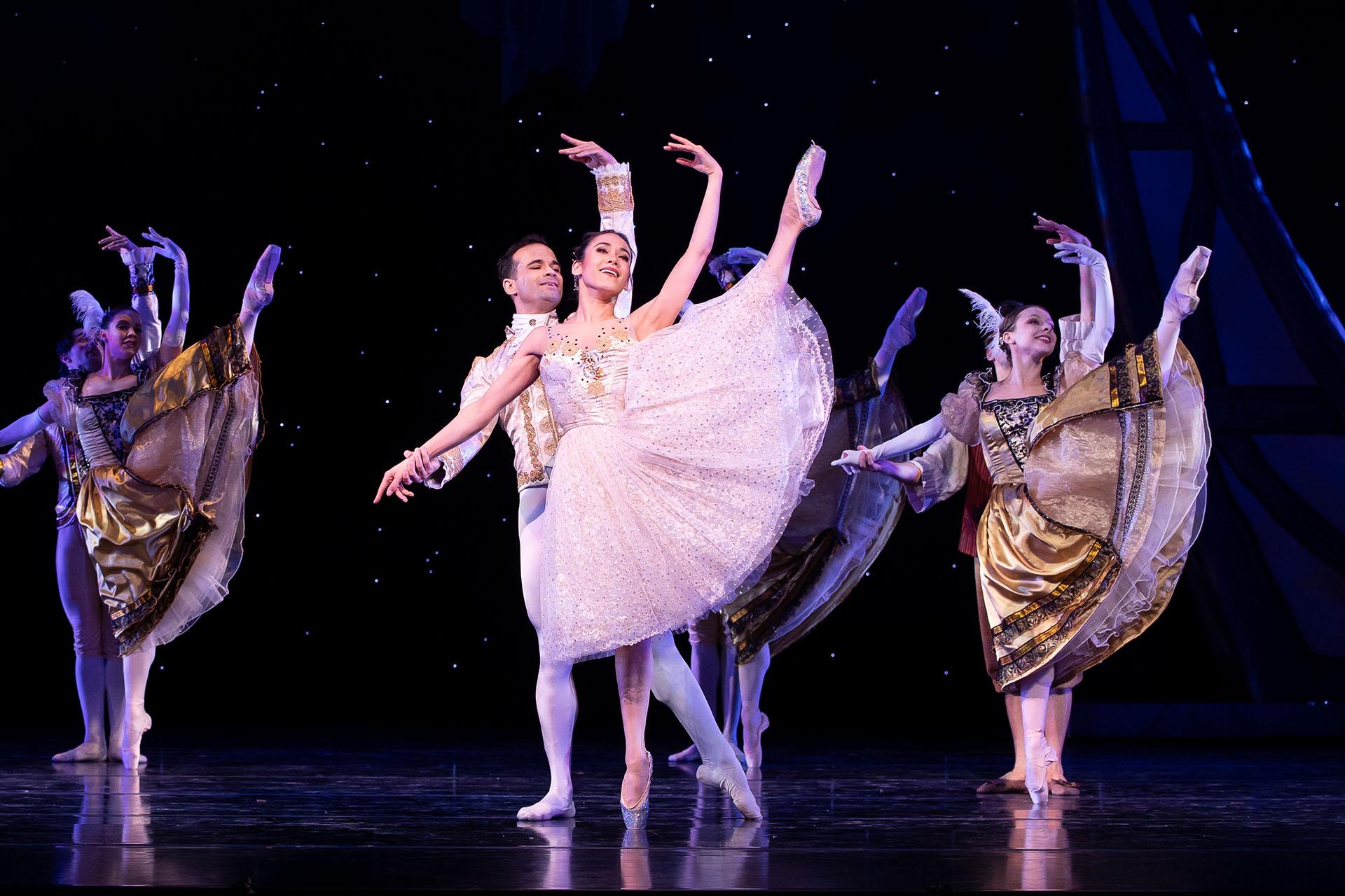 Woman in white-silver shimmering dress performs a grand battement while a regal man in white and gold supports her